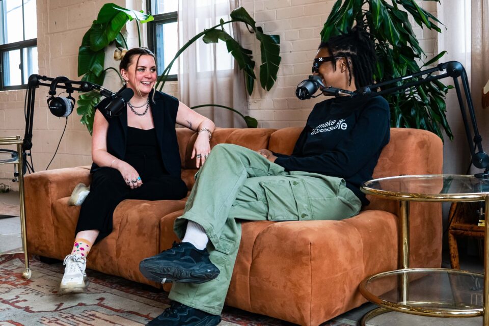 Two individuals are seated on a couch in a indoor setting. They are engaged in conversation, each speaking into microphones mounted on stands positioned in front of them.